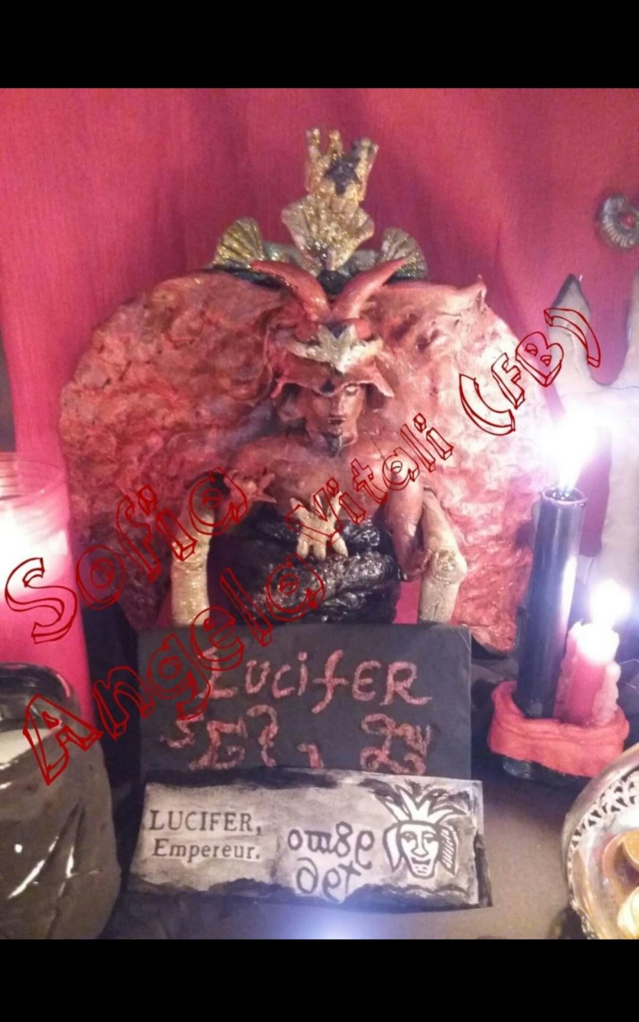 A close view of a sculptural display with a horned figure, carved plaque and candles, brightly lit against a red backdrop.
