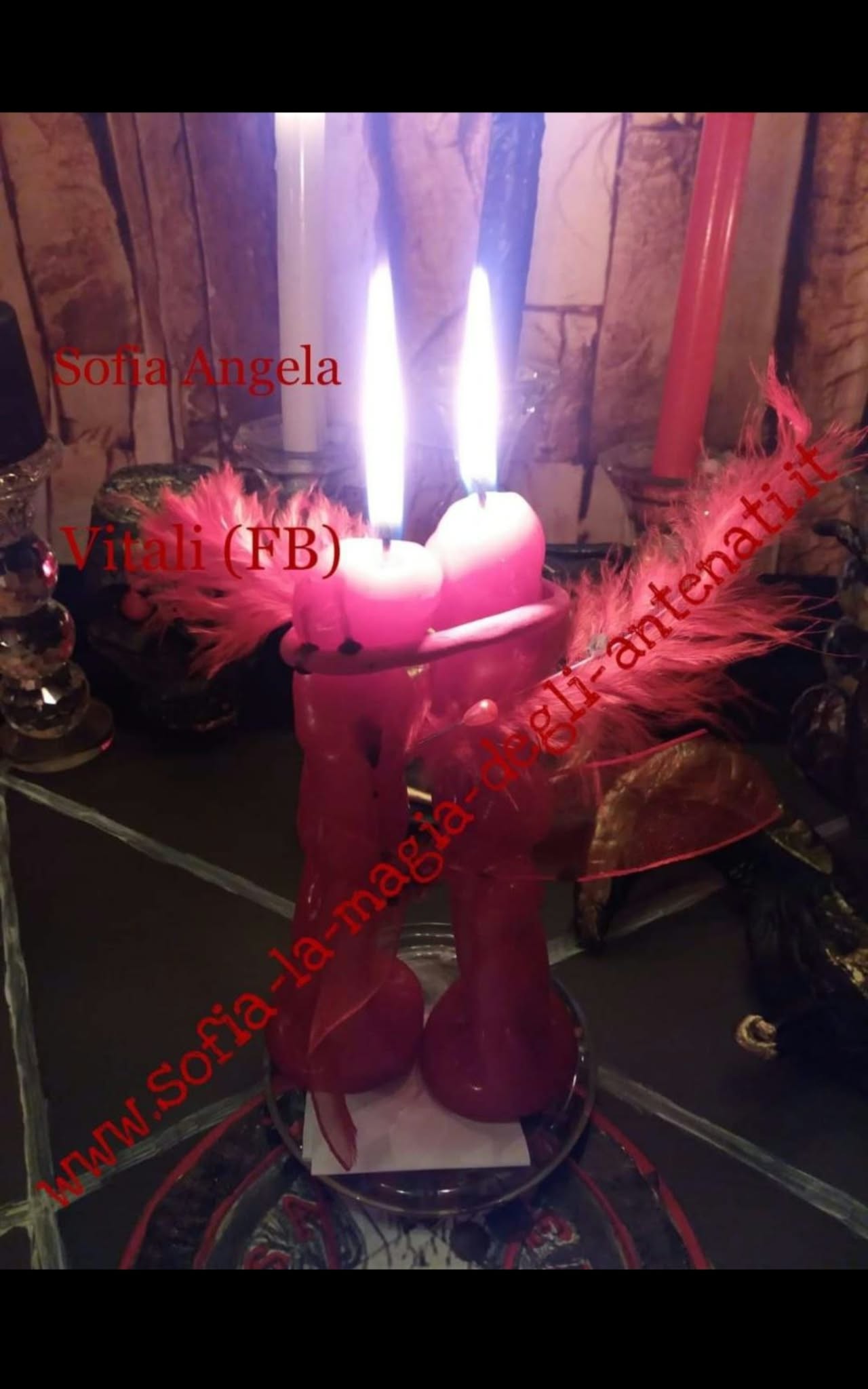 A close-up of two pink candles burning in a small holder, surrounded by red feathers and other objects in low light.
