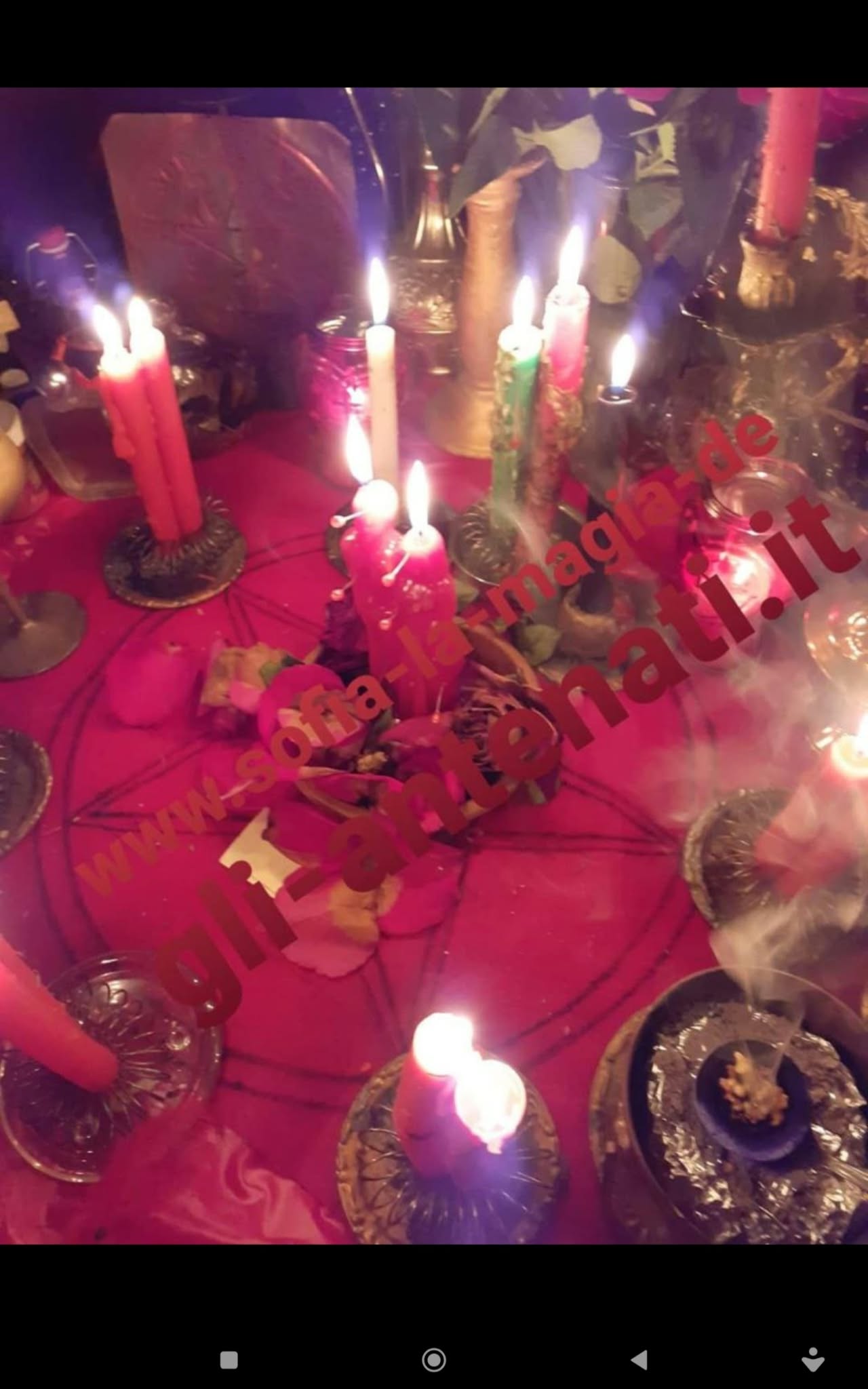 A close view of numerous lit red and pink candles, flowers and small objects arranged on a red cloth under warm light.